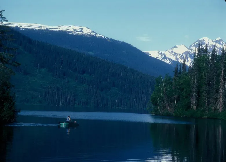 Meziadin Lake, an angler in a small boat below snow-capped peaks and dark forest