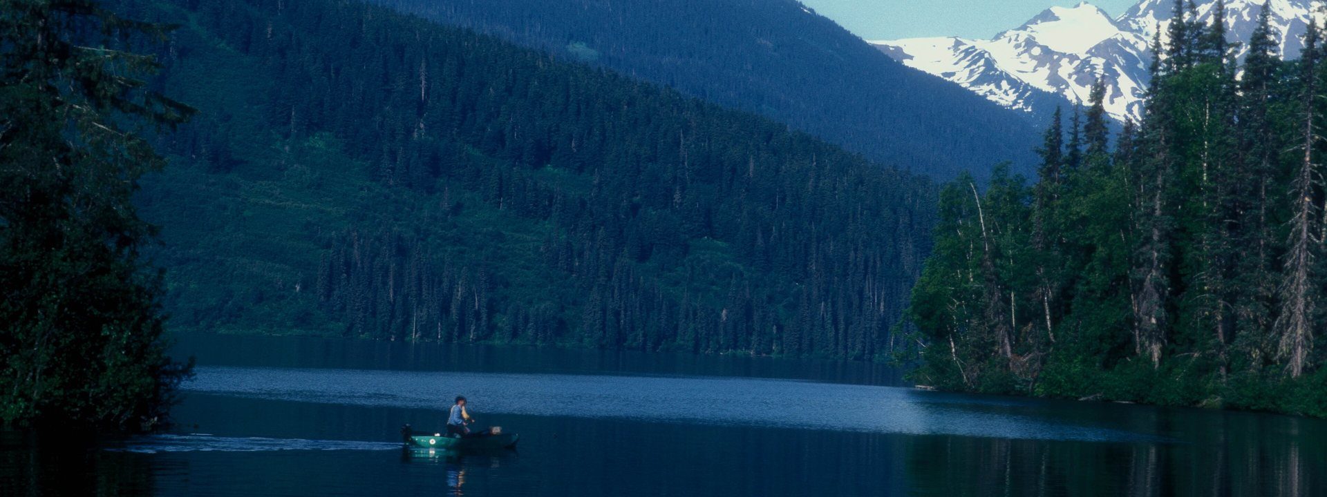 Meziadin Lake, an angler in a small boat below snow-capped peaks and dark forest